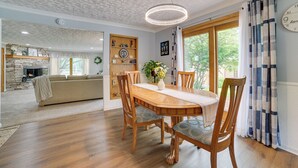 A dining area with a wooden table, chairs, and a chandelier. There is a fireplace in the background and a large window with curtains. - Northern Michigan Vacation Rental Near Trails! (Atlanta)
