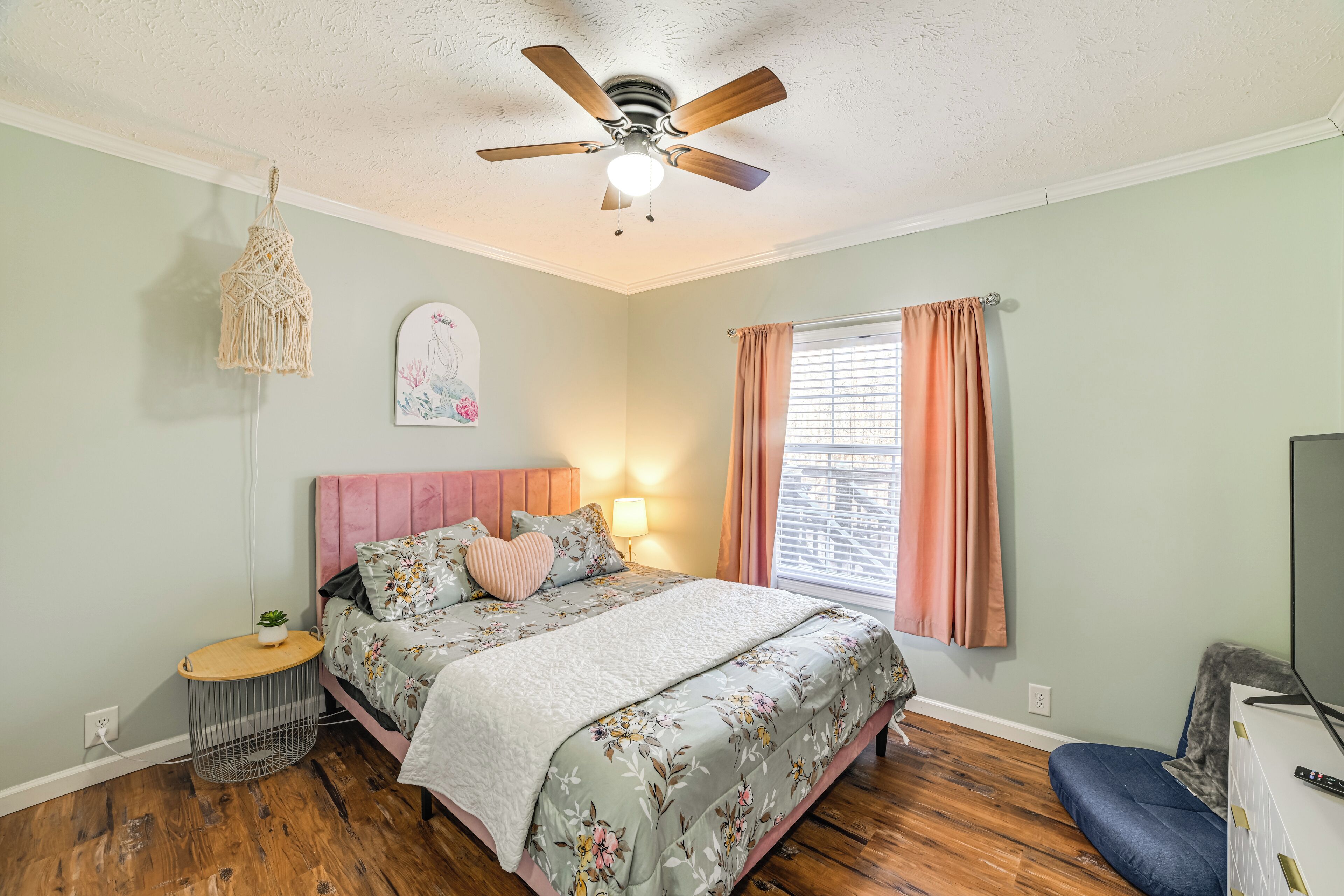 A bedroom with a bed, a ceiling fan, a window with curtains, a wall-mounted TV, and a small side table with a plant.