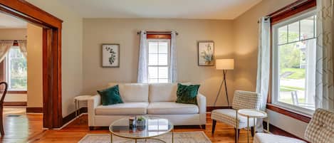A living room with a white sofa, a glass coffee table, a patterned rug, and a view of the outdoors through a window.
