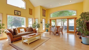 A spacious living room with a large sofa, a coffee table, and a dining area with a table and chairs. There are large windows and a view of trees outside. - 'lockes Glen' - Lakefront Geneva Getaway w/ Dock! (Geneva)