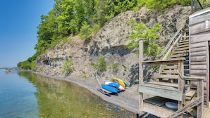 A wooden dock with a staircase leading to a platform, overlooking a calm body of water and a rocky cliff with sparse vegetation. - 'lockes Glen' - Lakefront Geneva Getaway w/ Dock! (Geneva)