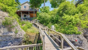A wooden staircase leading to a house with a balcony, surrounded by lush greenery and a rocky cliff. - 'lockes Glen' - Lakefront Geneva Getaway w/ Dock! (Geneva)