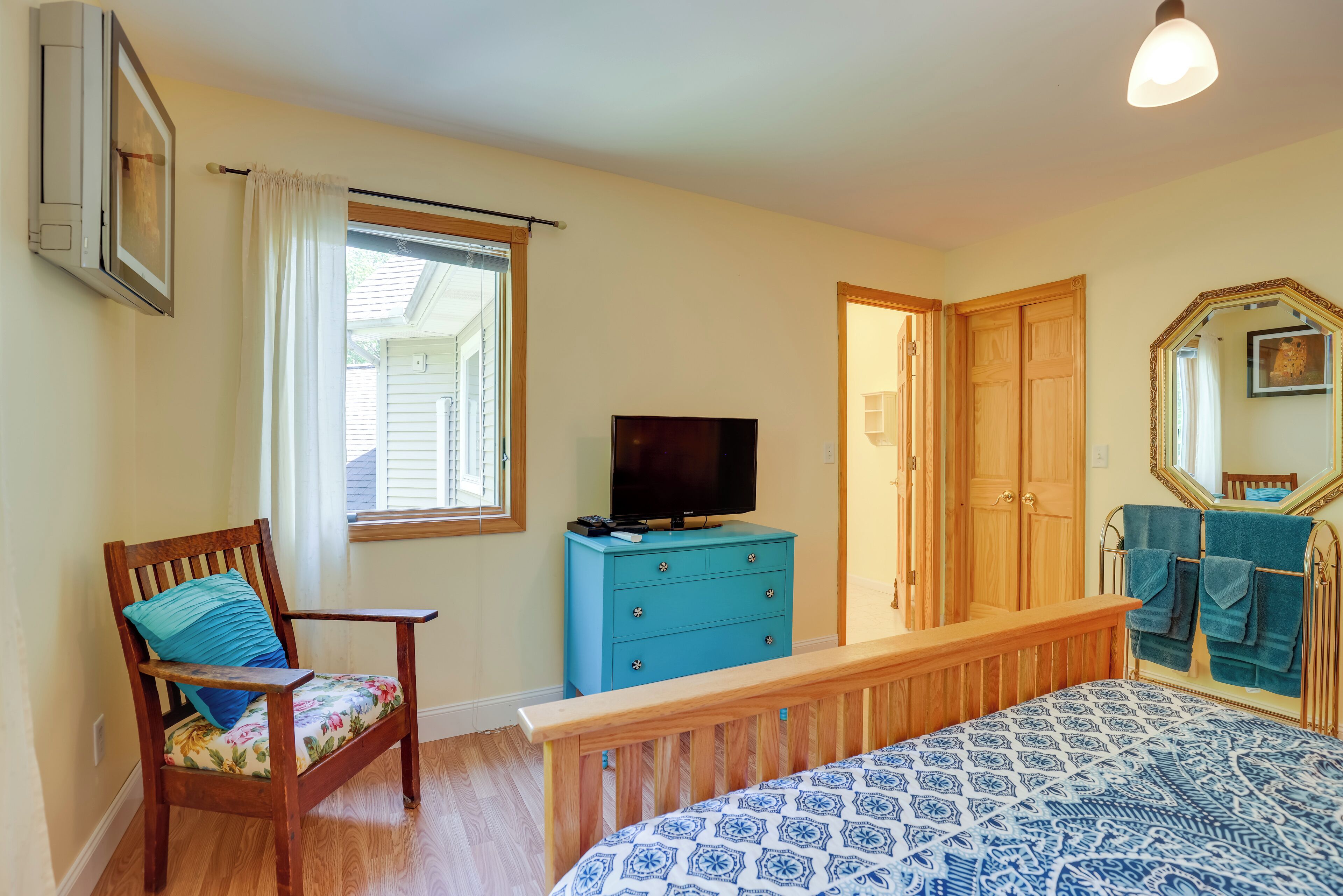 A bedroom with a wooden bed, a blue dresser, a television, a chair with a floral cushion, a window with white curtains, and a wooden wardrobe.
