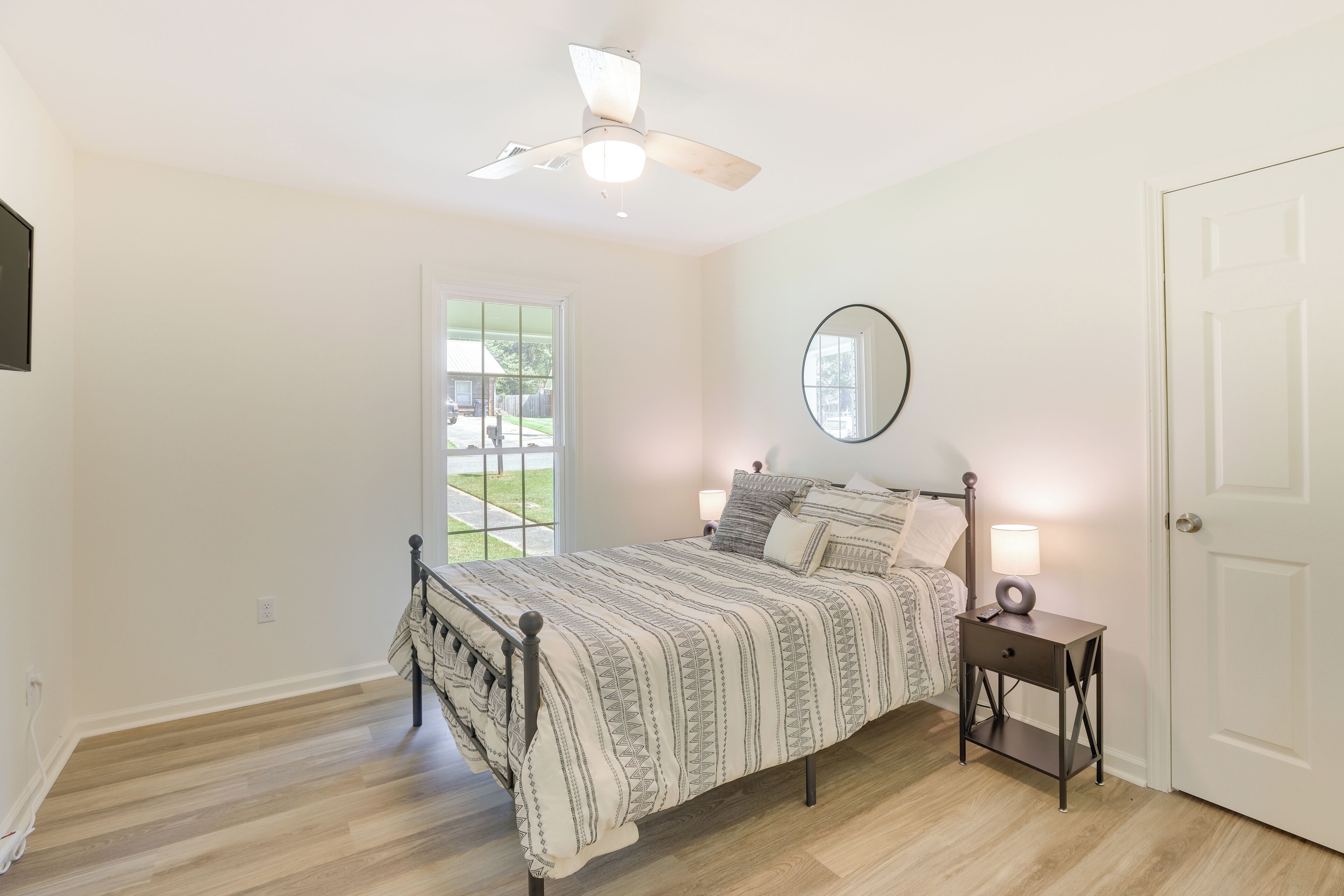 A bedroom with a bed, a bedside table with a lamp, a ceiling fan, a window, and a door.
