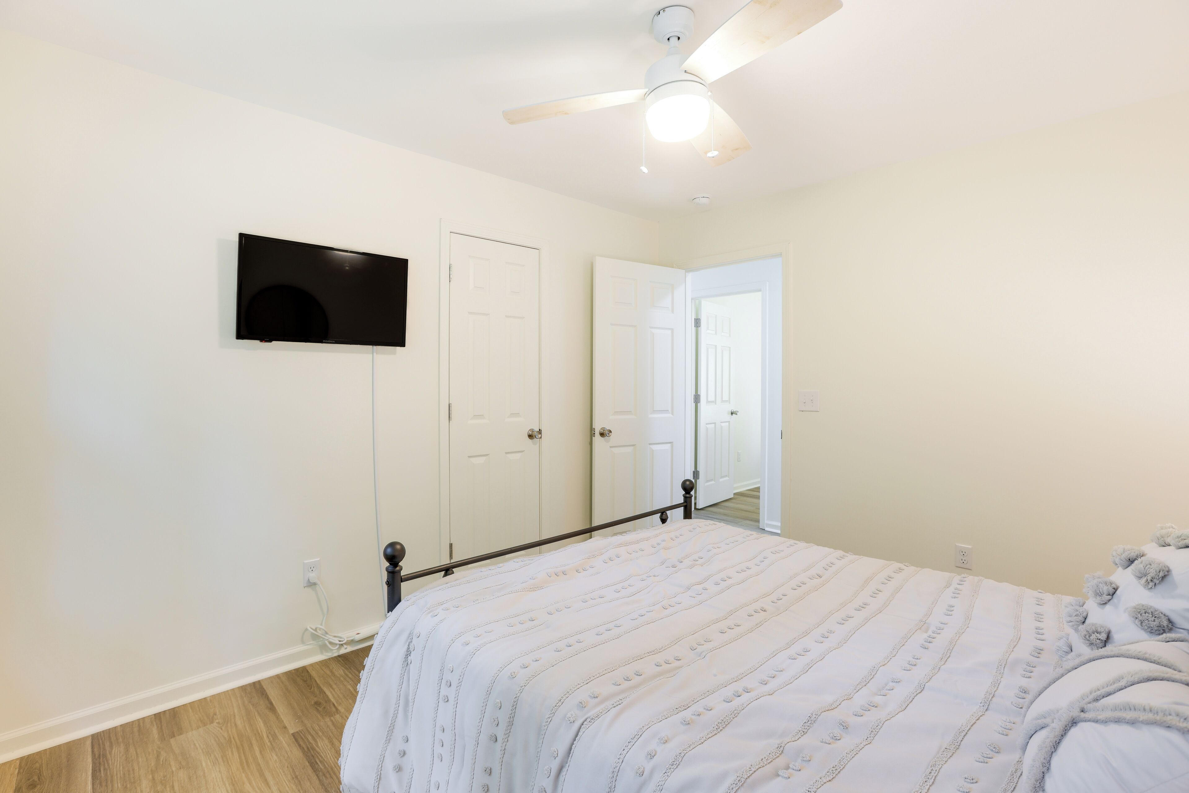 A bedroom with a bed, a ceiling fan, a TV mounted on the wall, and a door leading to another room.