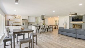 A modern kitchen with white cabinets, a central island, and bar stools. There is a dining area with a table and chairs, and a grey sofa in the living room. - Northport Game Day Getaway: 5 Mi to University! (Northport)