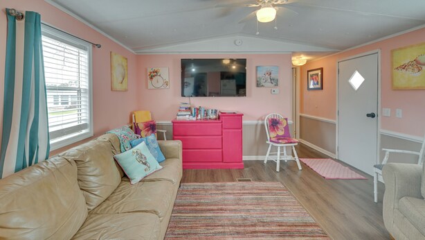 A living room with a beige sofa, a pink dresser, and a white chair. There is a ceiling fan, a window with curtains, and artwork on the walls.