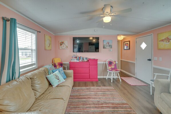 A living room with a beige sofa, a pink dresser, and a white chair. There is a ceiling fan, a window with curtains, and artwork on the walls.