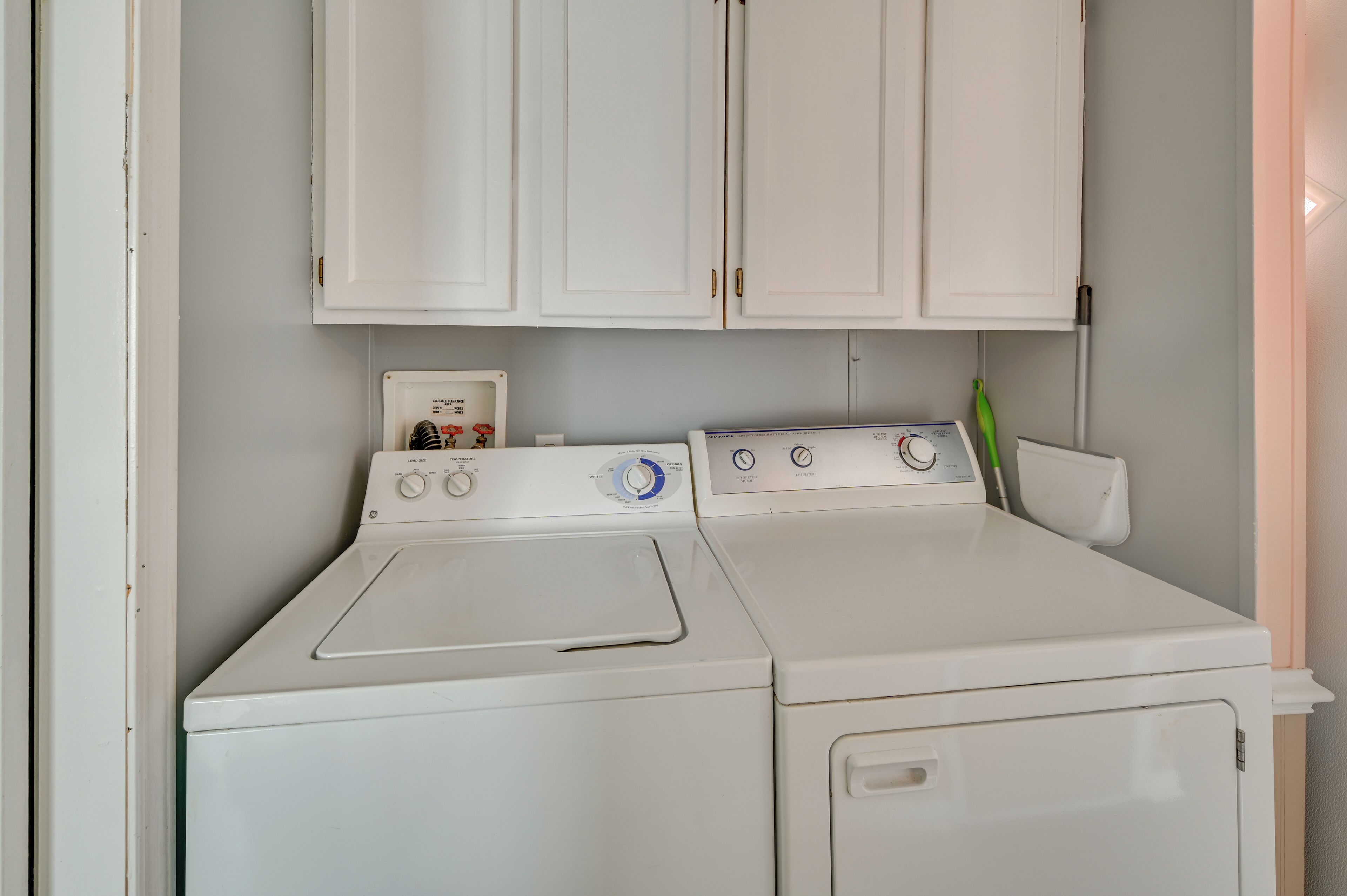 A laundry area with a front-loading washer and dryer, white cabinets above, and a green plant beside the dryer.