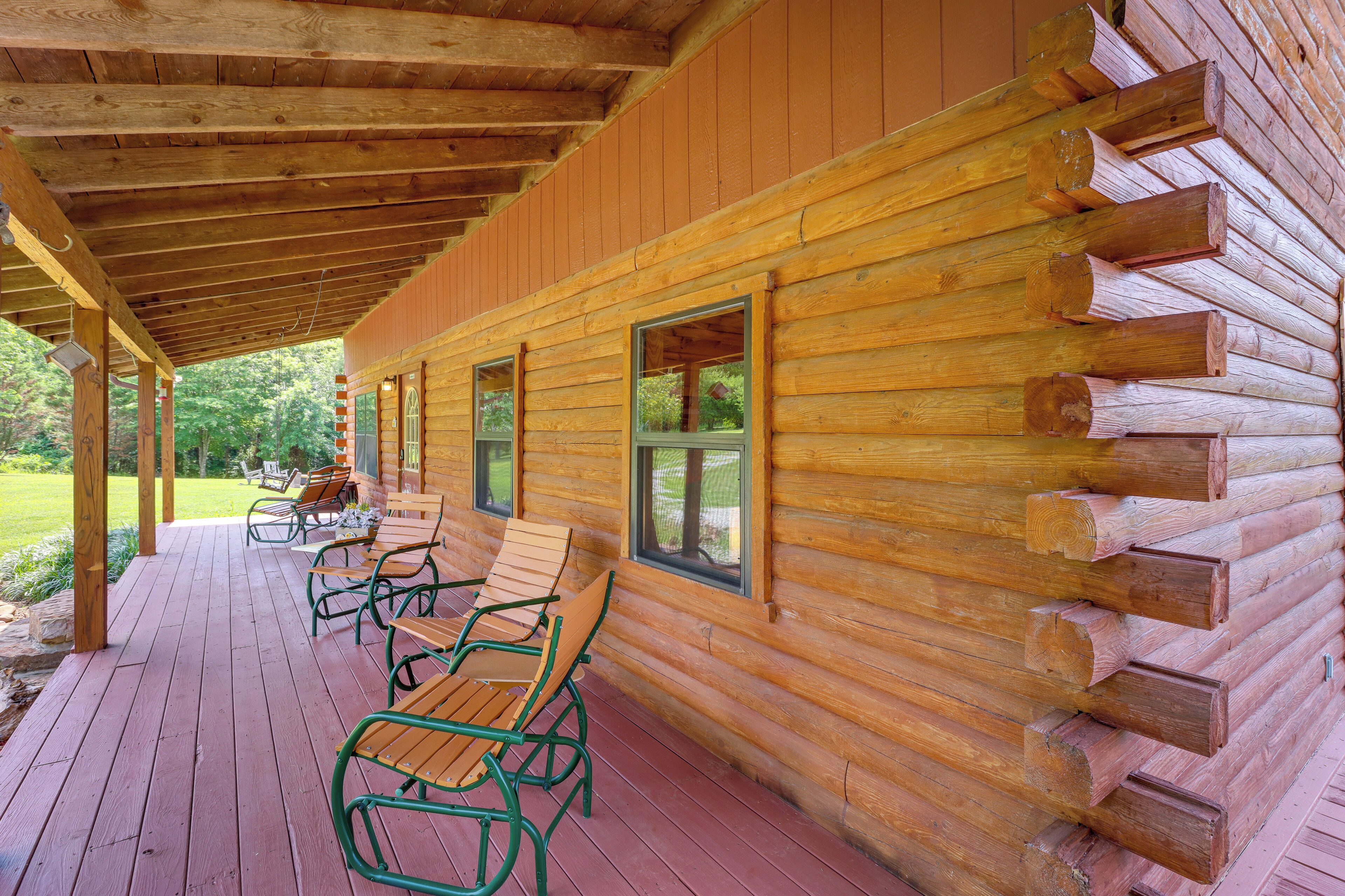 A log cabin with a covered porch, wooden benches, and a view of the surrounding forest.