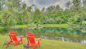 Two red Adirondack chairs by a calm lake surrounded by lush greenery. - Ambient Manchester Abode w/ Deck & Pond! (Manchester Center)