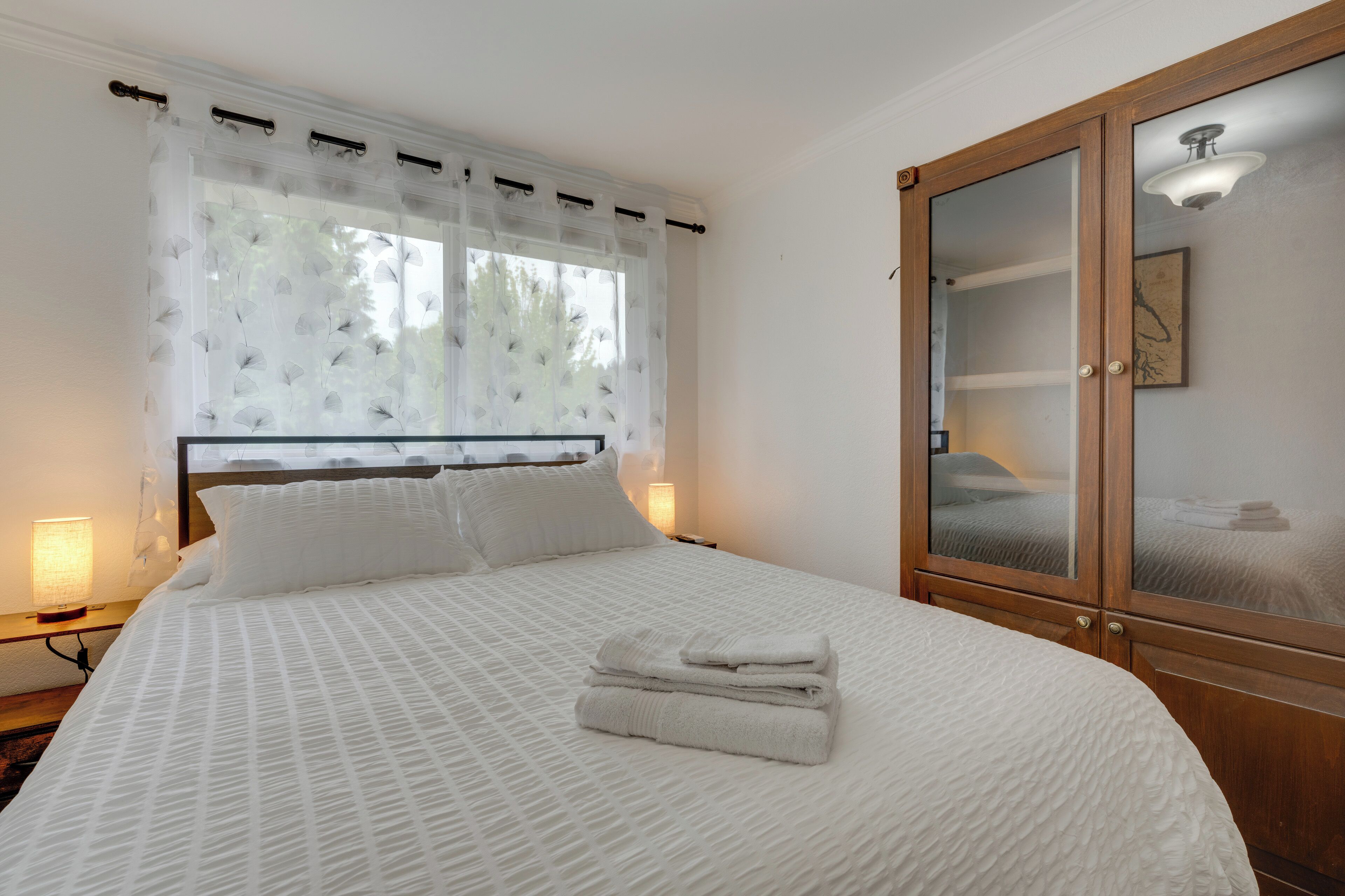 A neatly made bed with a white bedspread and pillows, a set of folded towels on the bed, and a wooden wardrobe with glass doors.
