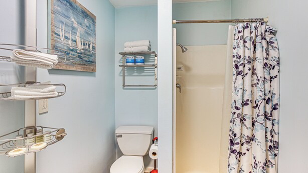 A bathroom with a toilet, a shower curtain, and shelves with towels and toiletries. - Walkable Paradise: Downtown Puyallup Studio! (Puyallup)