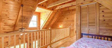 A wooden cabin room with a sloped ceiling, a bed, a window, and a door.