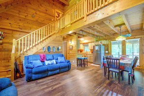 Interior de una cabaña con escalera de madera, un sofá azul, una mesa de comedor con sillas y una zona de cocina.