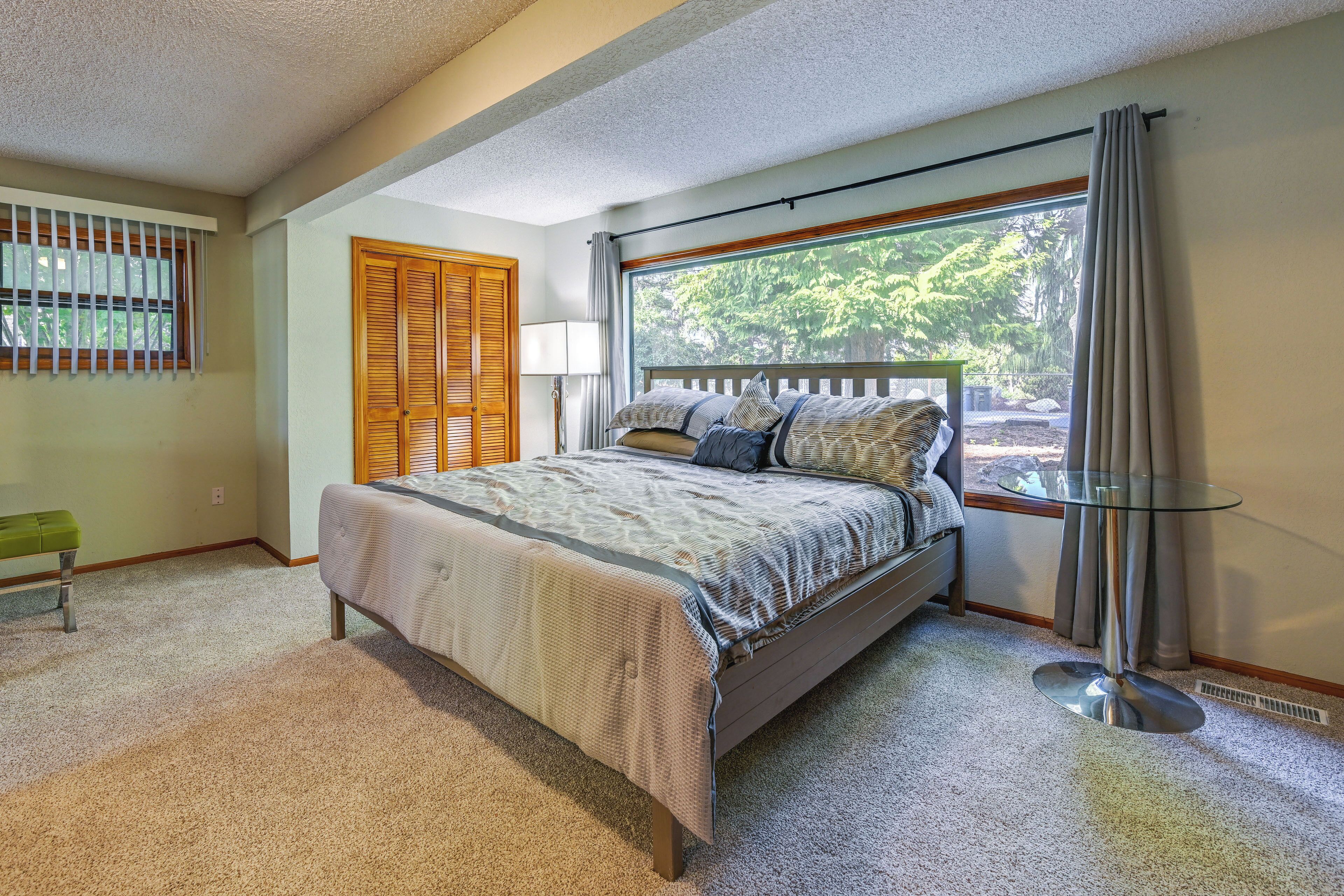 Quarto com cama, mesa de cabeceira, abajur, janela com cortinas e vista para árvores.
