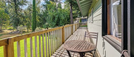 A wooden deck with a checkered floor, a table, and a chair, overlooking a grassy area with trees and a building in the background.