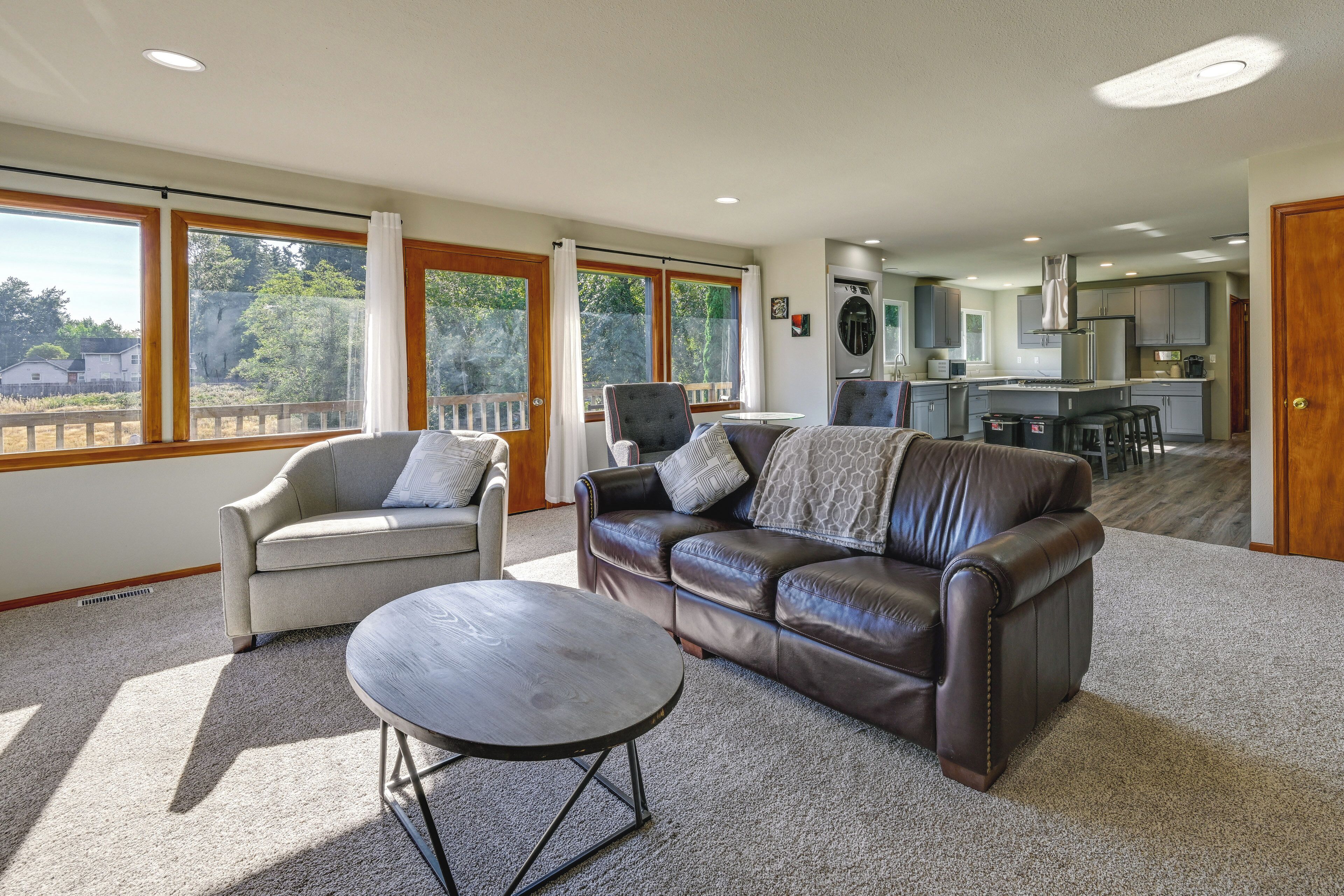 Ampla sala de estar com sofá de couro, poltronas e mesa de centro. Uma cozinha com eletrodomésticos modernos é visível ao fundo.