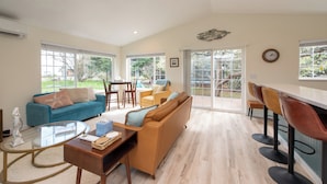 A spacious living area with a sofa, coffee table, and chairs. There is a dining table with chairs and a kitchen area with bar stools. Large windows provide natural light and views of the outdoors. - Sequim Escape w/ Gas Grill & Deck, Walk to Beach! (Sequim)