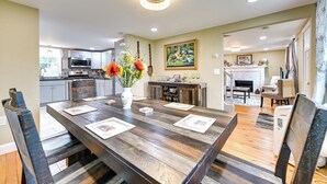 Un comedor con mesa de madera, sillas y un jarrón de flores. Se observa una cocina al fondo con microondas y armarios.