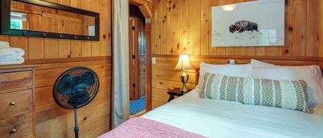 A wooden-paneled room with a bed, a standing fan, a mirror, and a wall-mounted picture.