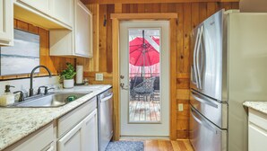A compact kitchen with white cabinets, a stainless steel refrigerator, a sink, and a door leading to an outdoor patio with a red umbrella. - Fire Pit, Deck & Grill: Cozy Twain Harte Cabin! (Twain Harte)