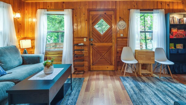 A cozy cabin interior with a wooden door, a sofa, a coffee table, a desk with chairs, and a bookshelf. - Fire Pit, Deck & Grill: Cozy Twain Harte Cabin! (Twain Harte)