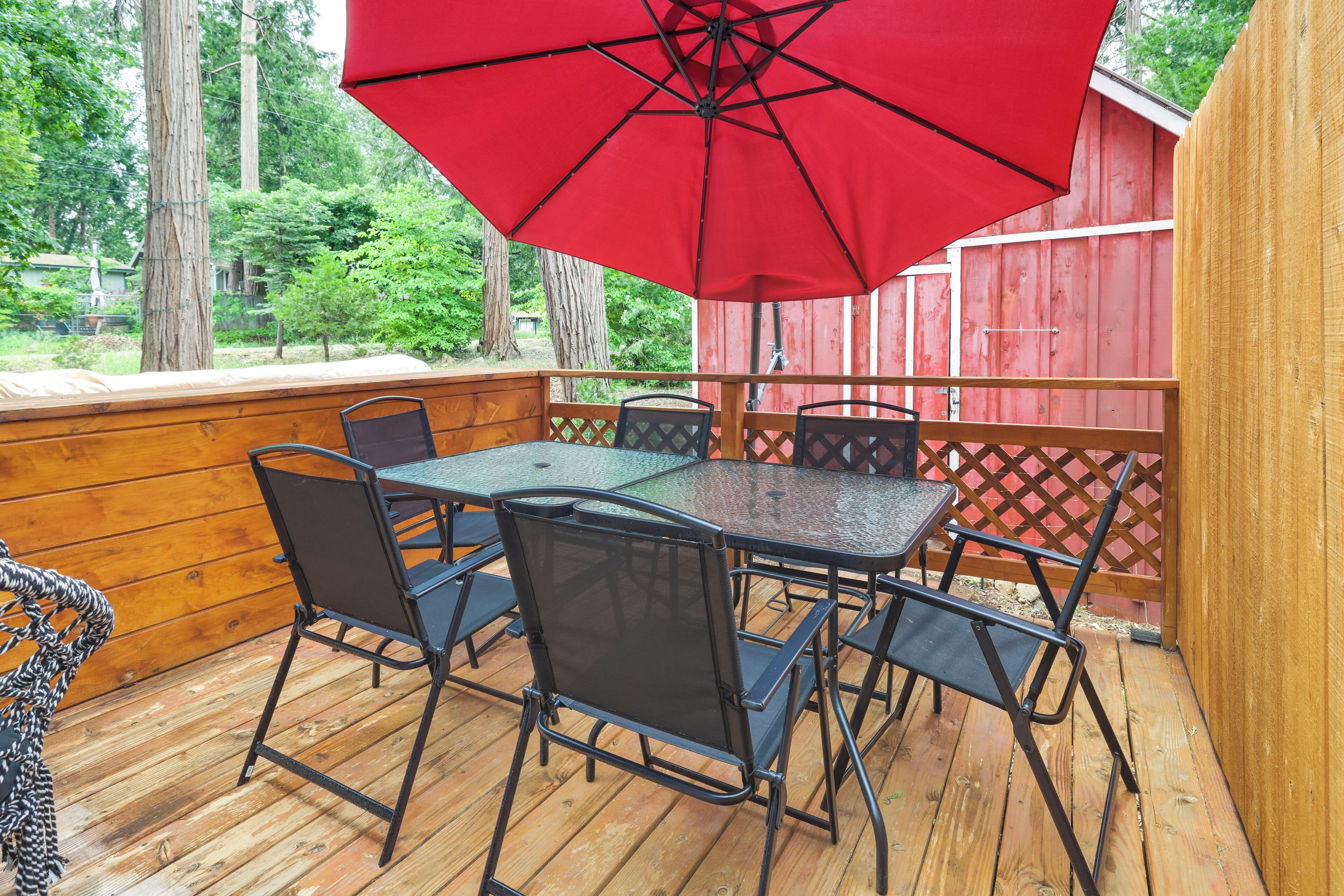 Une terrasse en bois avec une table à plateau de verre et quatre chaises, un parasol rouge et une clôture en bois en arrière-plan.