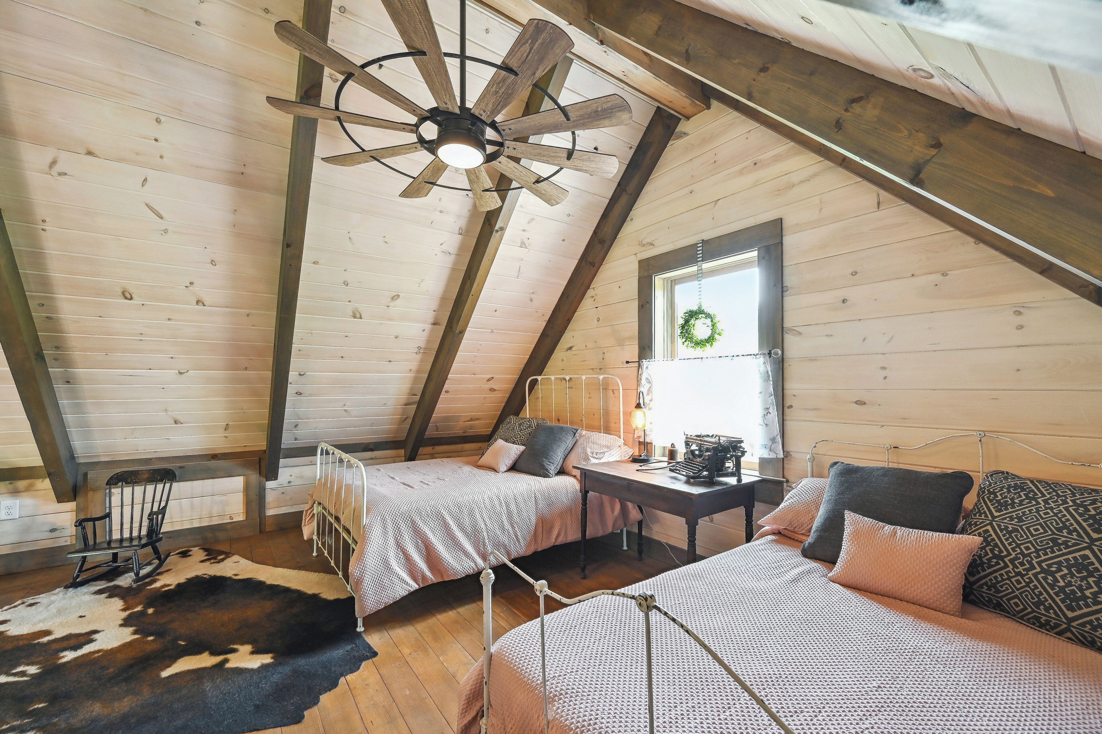 A cozy attic bedroom with two beds, a wooden ceiling fan, a desk, and a hanging light fixture.