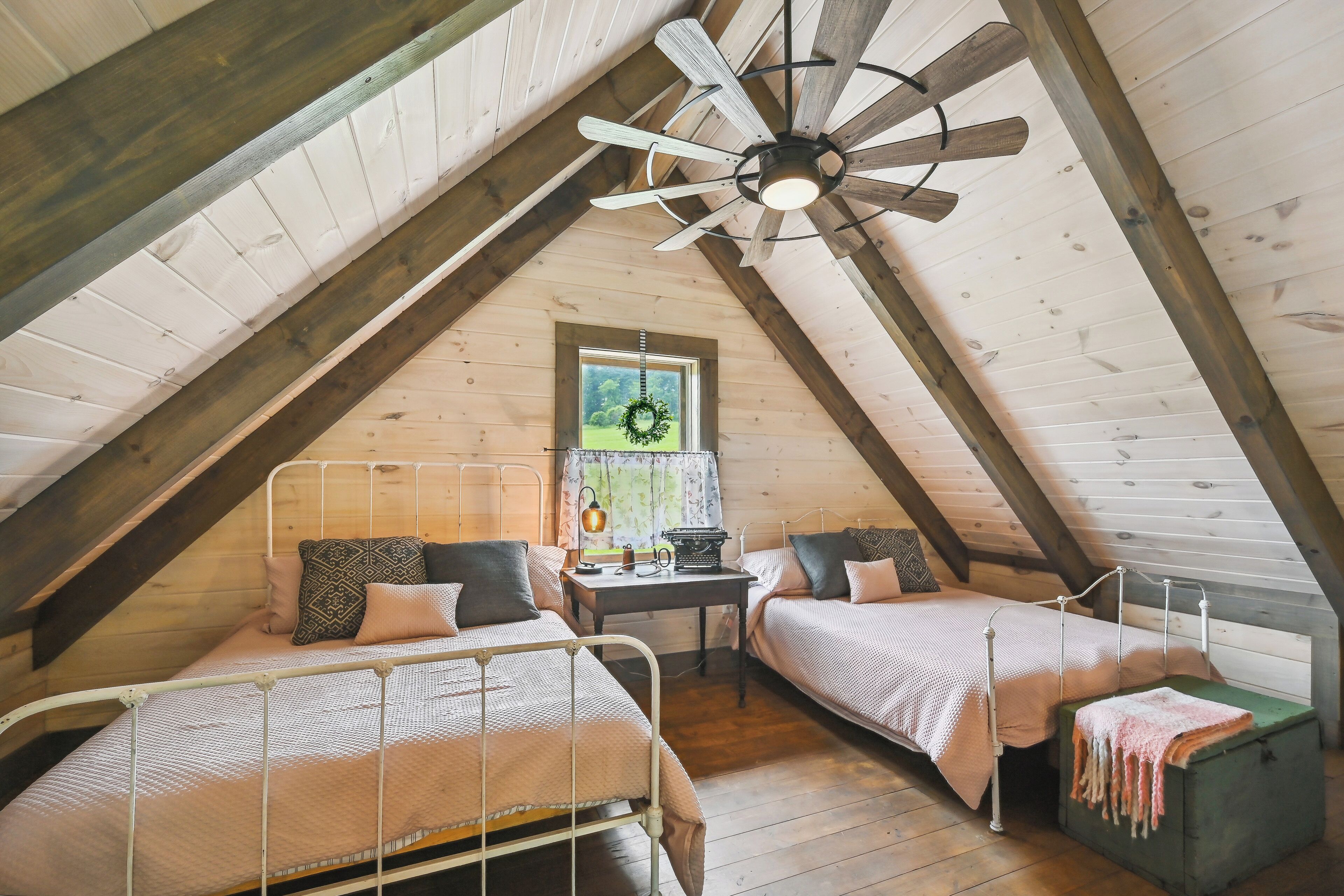 A cozy attic bedroom with two beds, a small table, and a window with a wreath.