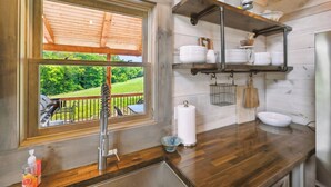 Interior de madera con una ventana que da a un paisaje verde, una encimera de cocina con un fregadero y estantes con vajilla y una percha.