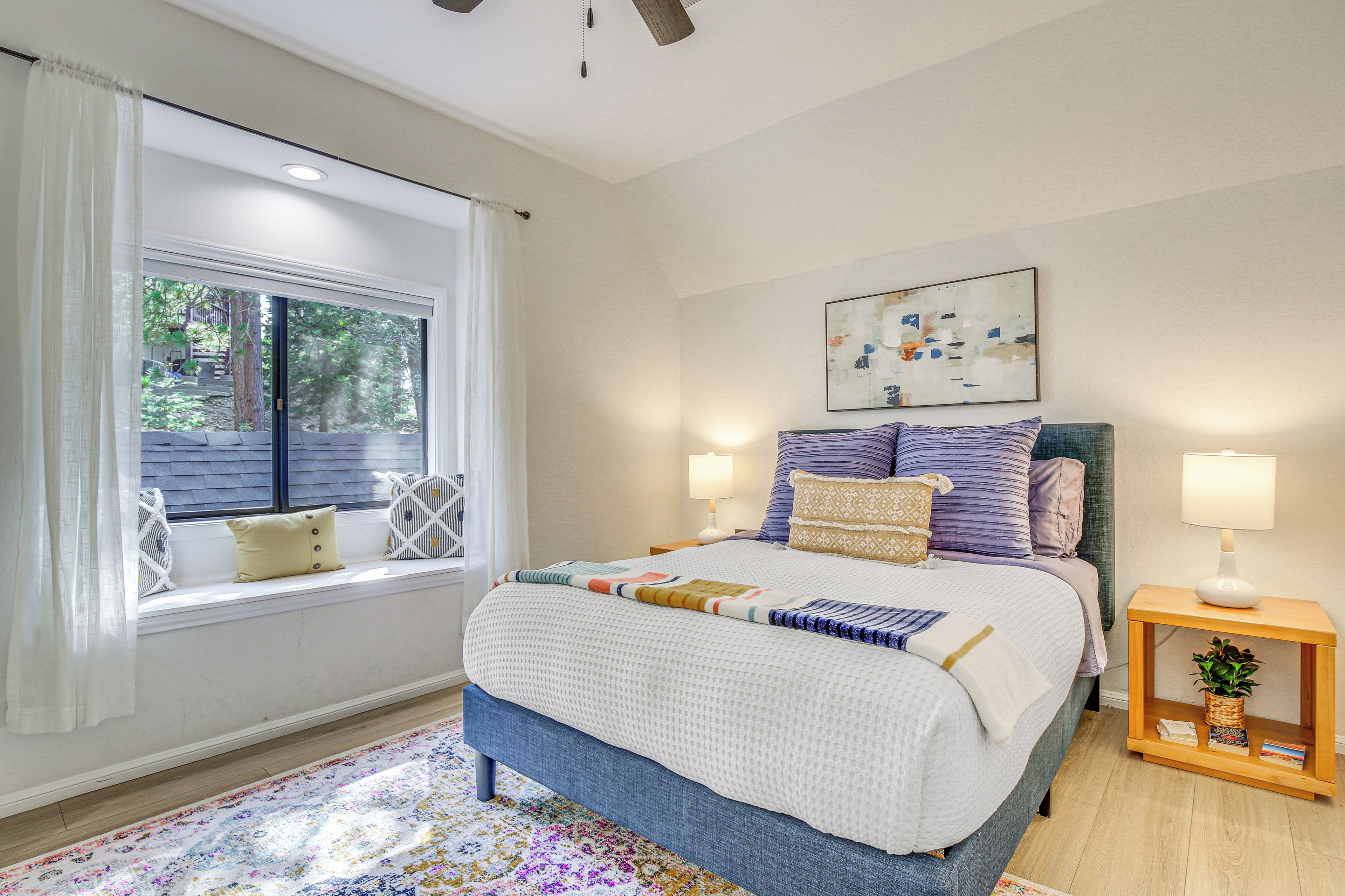 A bedroom with a large bed, a window with a view of trees, a ceiling fan, and a small bedside table with a lamp and a plant.