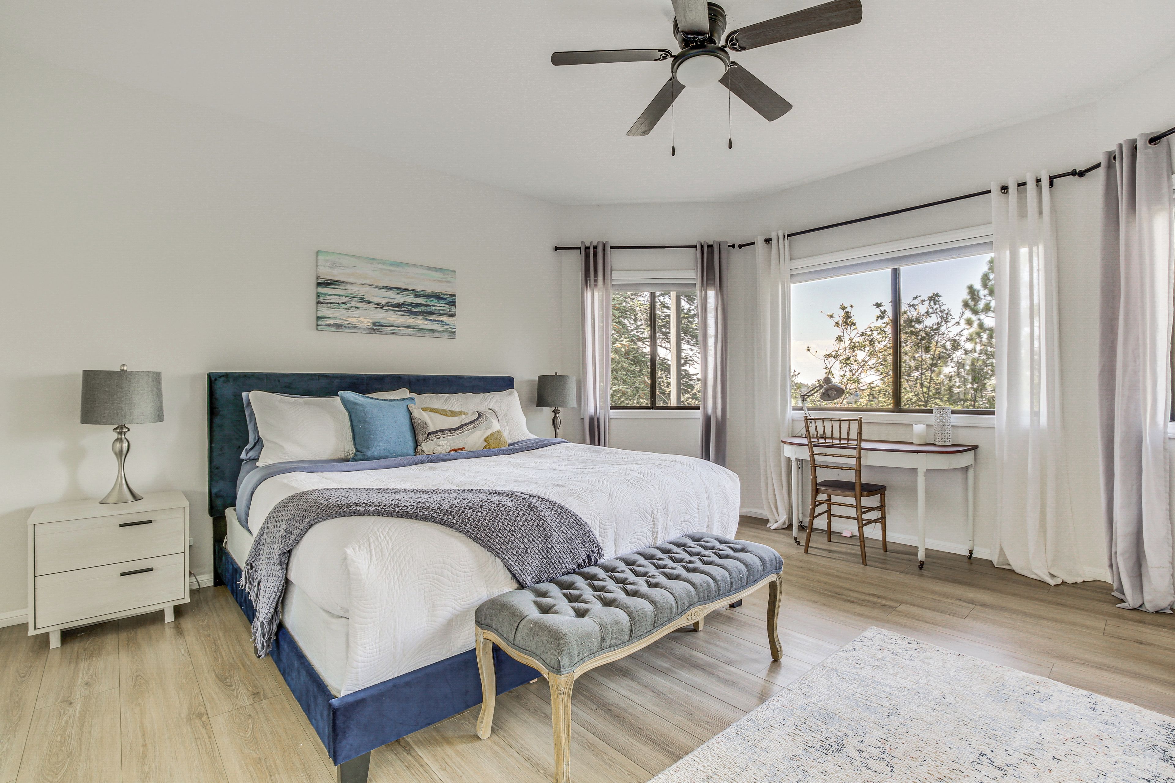 A bedroom with a bed, bedside table, lamp, bench, desk, and window with curtains.