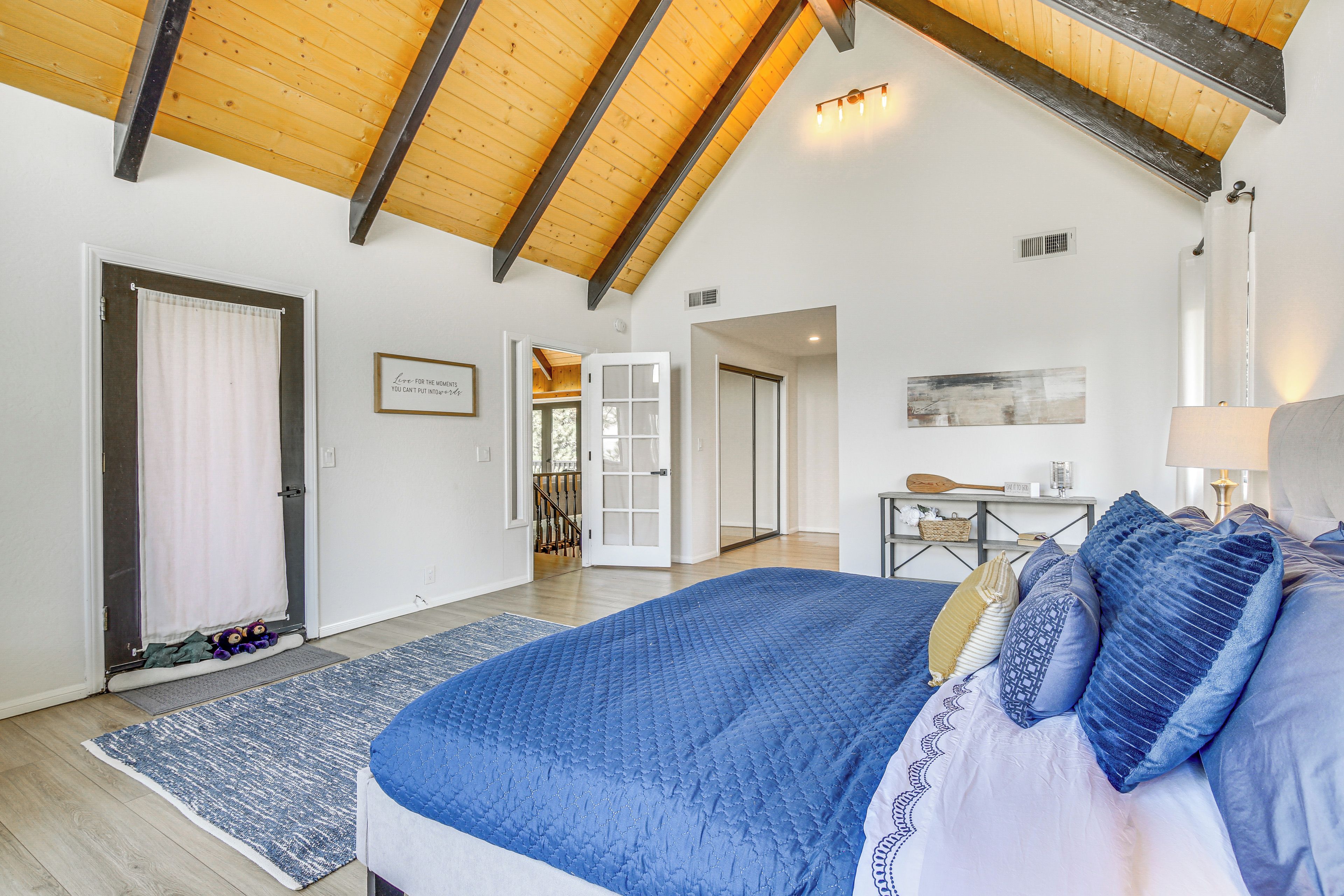 A bedroom with a large bed, wooden ceiling beams, a door with a curtain, and a small table with a lamp.