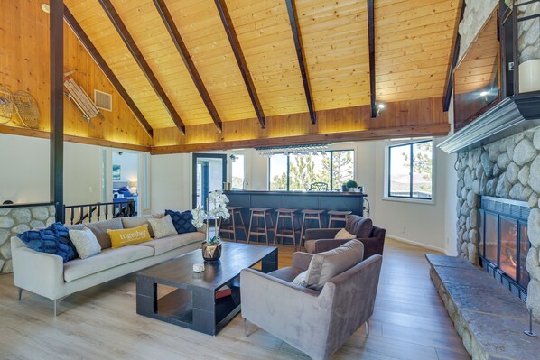 A spacious living area with a high wooden ceiling, a stone fireplace, and a modern kitchen in the background. - Lake Views & Game Room! Lake Arrowhead 3-king Gem (Lake Arrowhead)