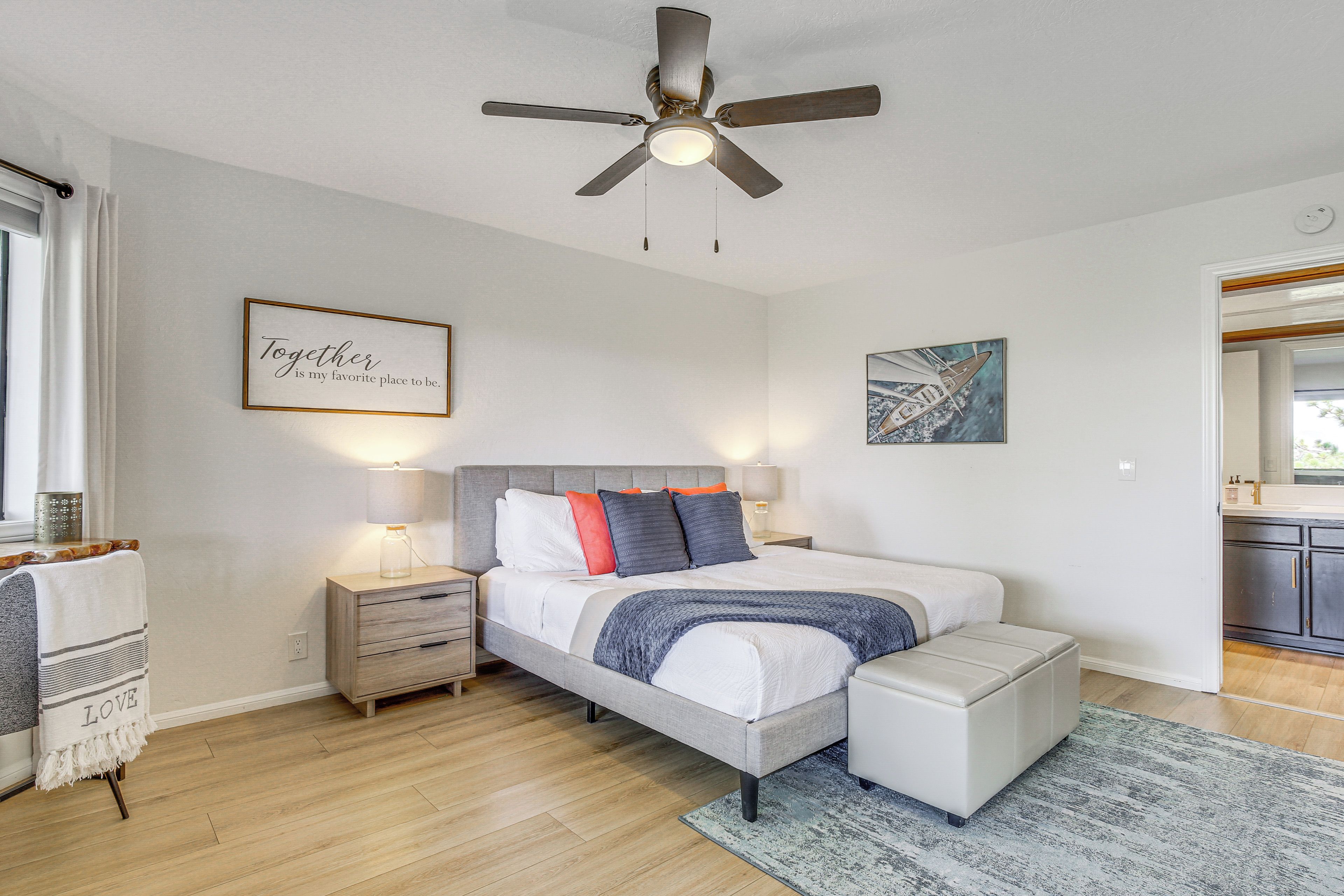 Un dormitorio con cama, mesitas de noche, un ventanal con cortinas y una cocina visible a través de una puerta abierta.