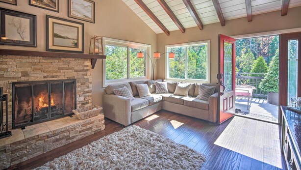 A cozy living room with a stone fireplace, a sectional sofa, and large windows offering a view of the outdoors. - Tree-lined Twin Peaks Cottage w/ Custom Stream (Lake Arrowhead)