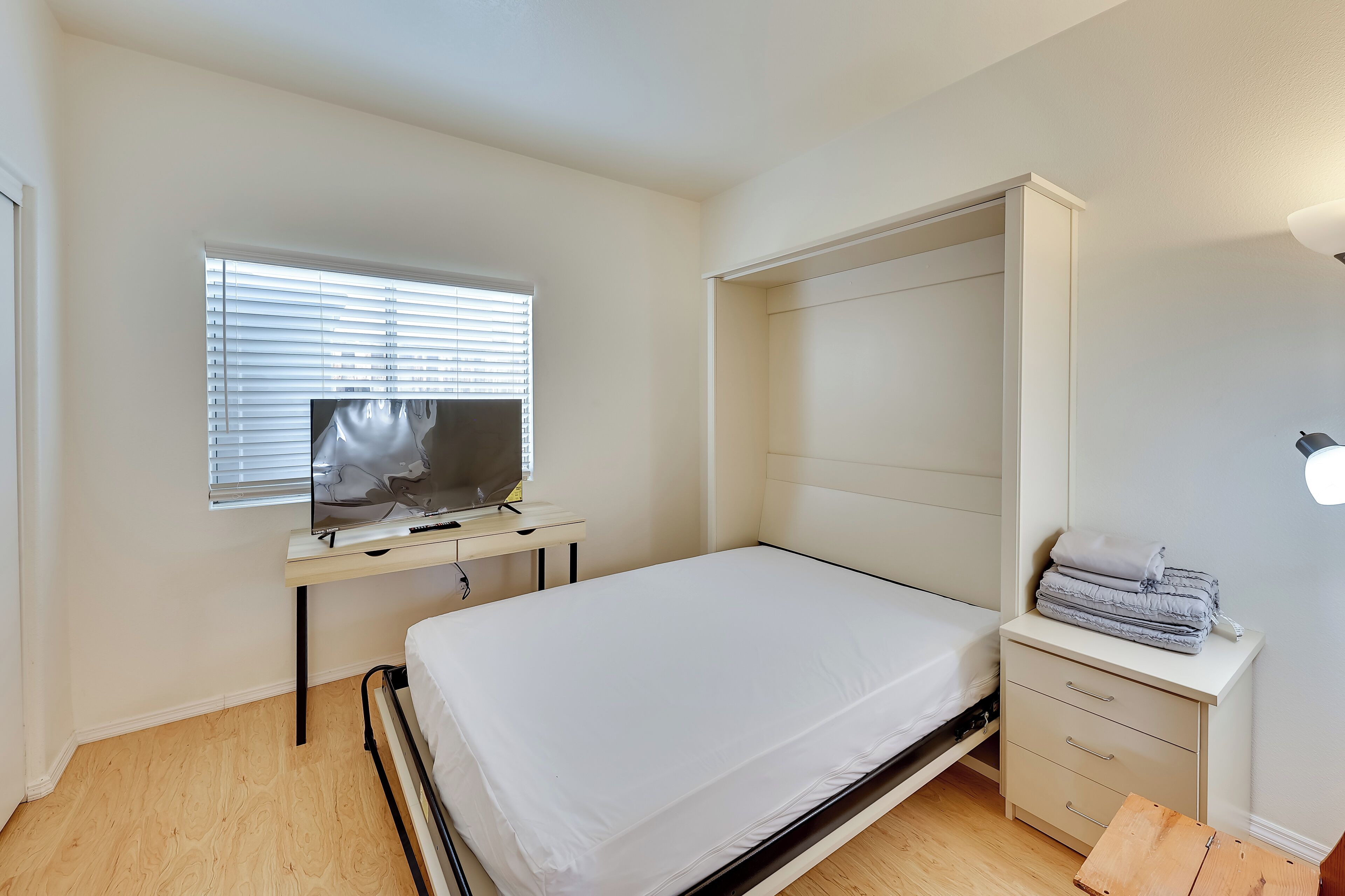 A bedroom with a bed, a desk, a wardrobe, and a window with blinds.