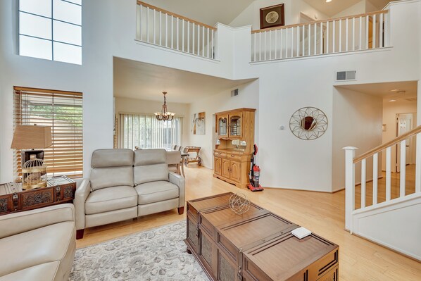 A spacious living area with a high ceiling, a large window, a beige sofa, a wooden coffee table, and a staircase leading to an upper level.