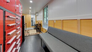 Une pièce de taille modeste, avec un canapé gris, un meuble de rangement rouge, une petite table avec des chaises et une fenêtre équipée de stores.