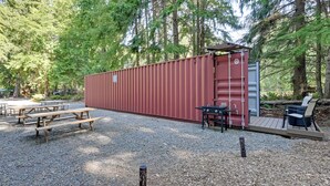 Un bâtiment de couleur rouge, de type conteneur, doté d’une terrasse et de tables de pique-nique, situé dans une zone boisée.