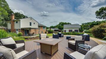 An outdoor deck with wicker furniture and a central table, surrounded by lush greenery and residential buildings.