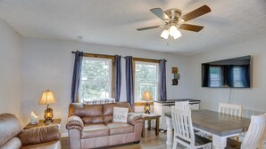 A furnished living room with a sofa, a dining table with chairs, a ceiling fan, and a window with curtains.
