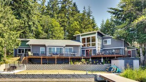A modern house with a deck overlooking a body of water, surrounded by dense forest. - Gorgeous Long Lake Home w/ Dock, Kayak, SUP & More (Lacey)
