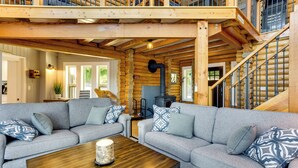 A log cabin interior with a wooden staircase, two grey sofas with patterned cushions, a wooden coffee table, and a fireplace. - Gorgeous Long Lake Home w/ Dock, Kayak, SUP & More (Lacey)