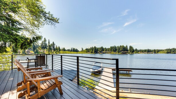 A wooden deck with outdoor furniture overlooking a lake, surrounded by trees and a clear sky. - Gorgeous Long Lake Home w/ Dock, Kayak, SUP & More (Lacey)