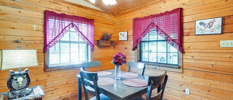 A wooden cabin interior with a dining table, chairs, a chandelier, and windows with purple curtains.
