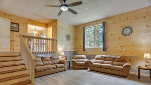 Un intérieur en bois avec un escalier, deux canapés en cuir, un ventilateur de plafond et une fenêtre donnant sur la forêt.