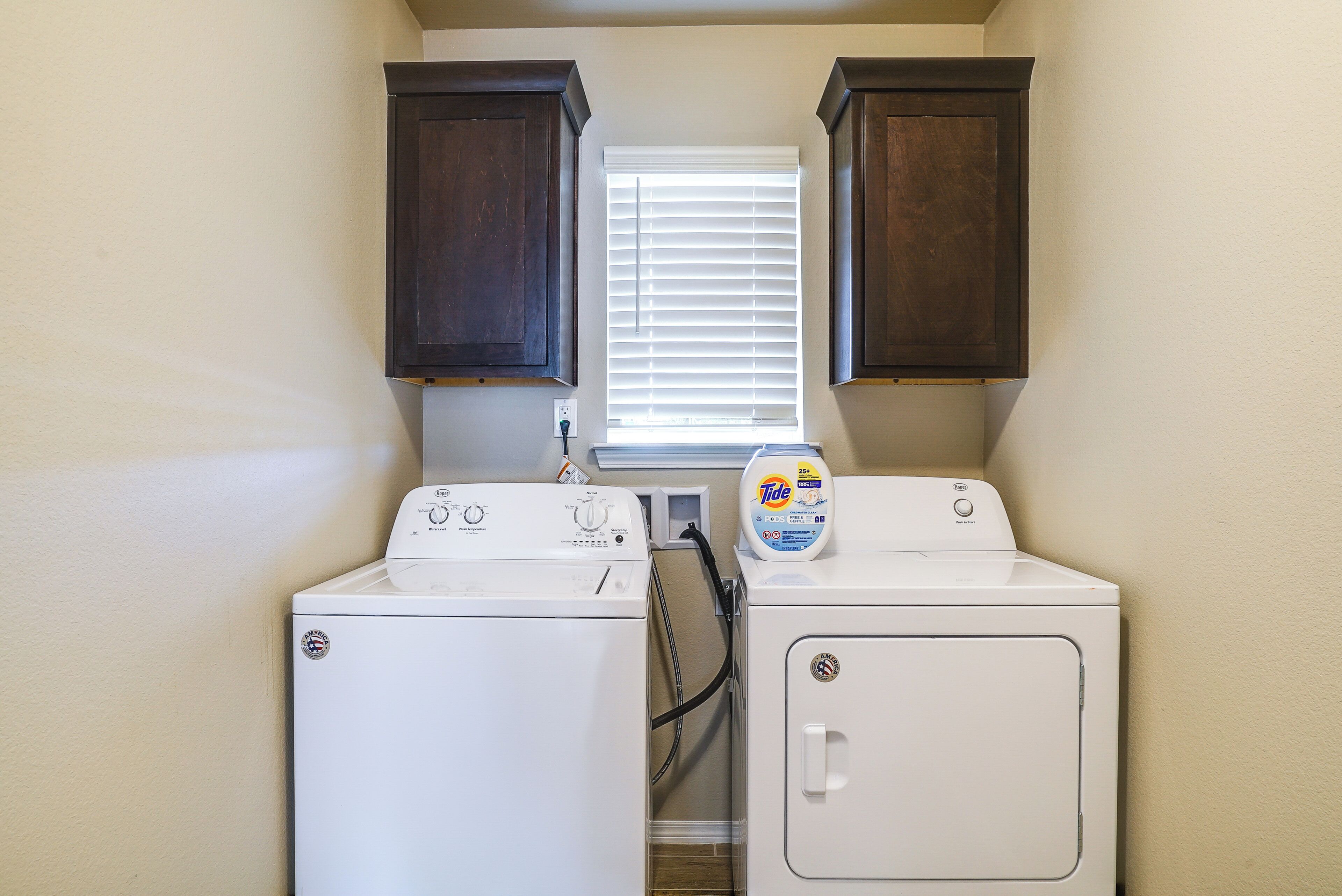 A laundry area with two washers and dryers, wooden cabinets above, and a window with blinds.