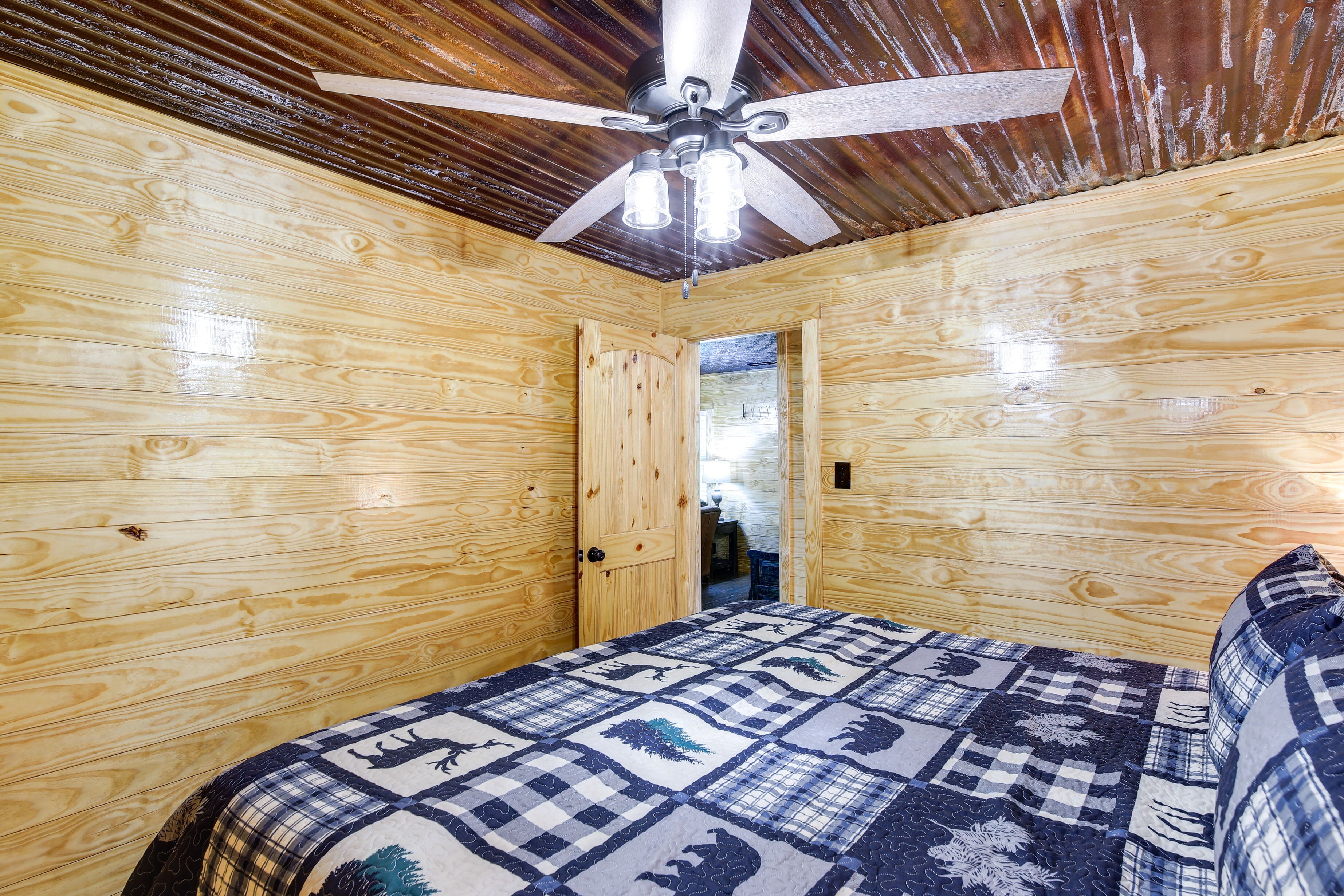 A wooden room with a bed, a ceiling fan, and a door leading to another room.